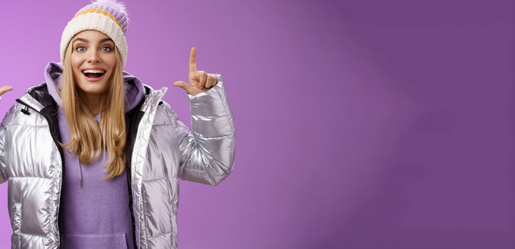 Excited carefree cheerful fair-haired european girl in silver jacket winter hat raising hands pointing up have excellent idea smiling broadly speaking passionately standing purple background photo