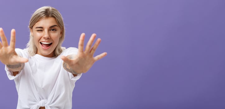 Reaching to you. Portrait of flirty good-looking and confident stylish woman with tattoos on arms winking sticking out tongue playfully and smiling pulling hands towards camera over purple background photo
