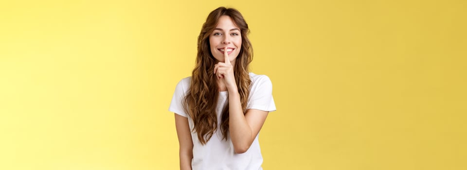 Cute cunning lovely european girl curly hairstyle hiding beauty secret smiling sensually show hush shush gesture index finger pressed lips grinning joyfully stand yellow background photo