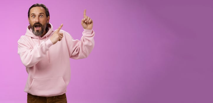 Impressed shocked exctied cool hipster mature 50s man bearded grey hair in pink hoodie retelling incredible story pointing up index fingers widen eyes surprised thrilled awesome advertisement photo