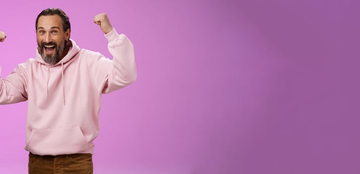 Yeah who cares age. Carefree delighted happy old man bearded in trendy pink hoodie raise fists joyfully triumphing having fun glad win celebrating success accomplish goal, posing purple background photo
