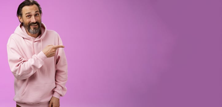 Friendly carefree caucasian bearded man in pink hoodie pointing right intoducing family members smiling proudly delighted showing blank copy space advertising, posing purple background photo