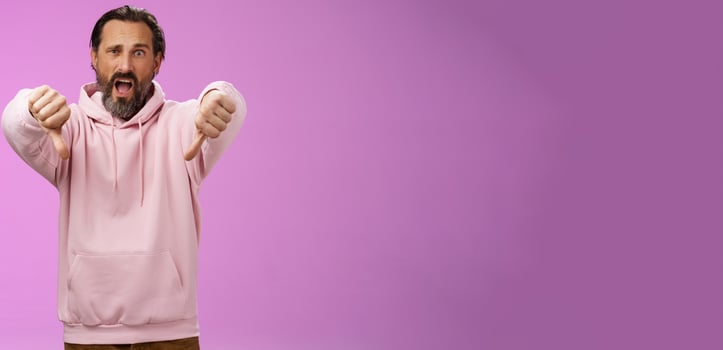 Extremely disappointed furious male customer swearing show thumbs down impulsive gesture yelling displeased pissed express dislike disgust look judgemental bothered, standing purple background photo