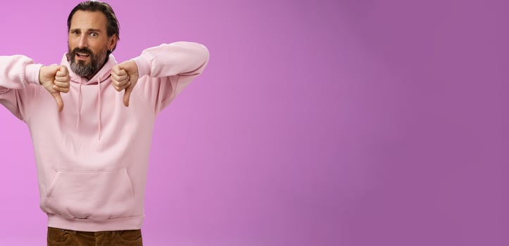 Disappointed upset angry caucasian adult bearded male show thumbs down dislking bad unprofessional behaviour frowning demanding refund standing bothered give negative feedback, purple background photo