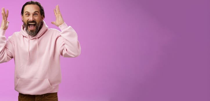 Happy excited lucky adult bearded man celebrating excellent news raising hands gesturing thrilled smiling broaldy triumphing victory win, yelling gladly achieve goal, standing purple background photo