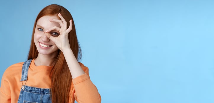 Girl search perfection. Charming pretty glad redhead female student assuring everything ok showing okay sign eye look through smiling delighted express positive attitude, blue background photo