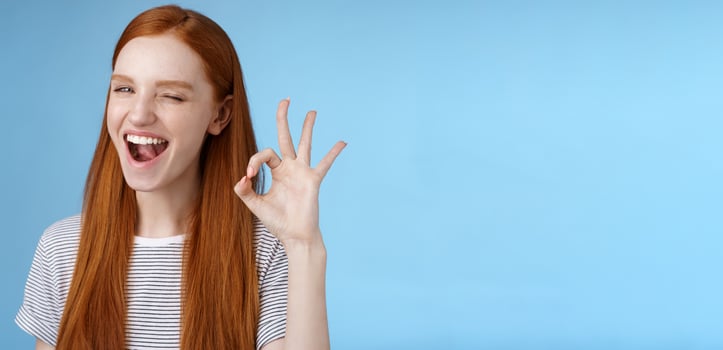 Sassy attractive redhead girl winking mysteriously smiling broadly give approval sign show okay ok excellent gesture satisfied good choice agree great decision, standing delighted blue background photo