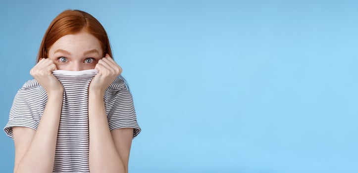 You cannot see me. Silly cute young teenage redhead girl hiding face pulling t-shirt head peeking look excited surprised camera hold collar standing funny under disguise, blue background photo