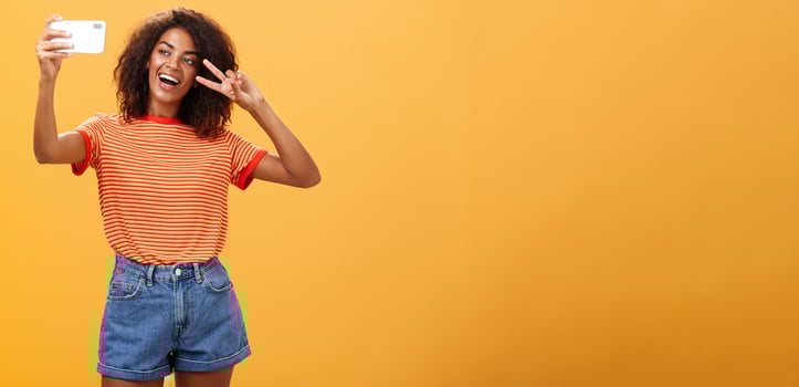 Girl making video vlog with brand new smartphone posting in internet trying become famous standing over orange background posing for selfie gazing at gadget screen showing peace or victory gesture photo