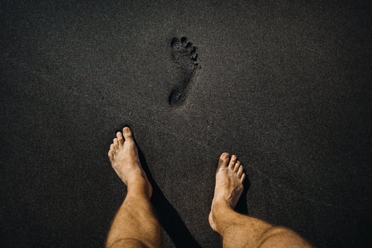 Close up of male footprints and feet walking on the volcanic black sand on the beach photo
