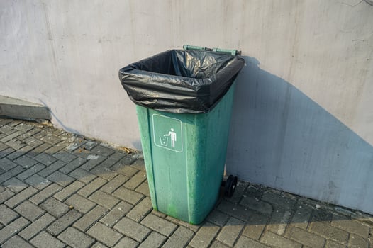 a green trash can with a black plastic trash bag inside on a paving block next to a white wall photo
