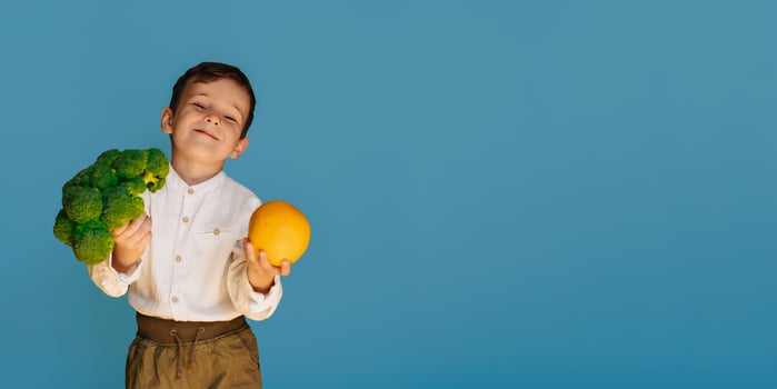 A studio shot of a boy holding fresh broccoli and an orange on a blue background with a copy of the space. The concept of healthy baby food. photo
