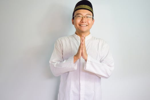 Asian Muslim man wearing glasses and white cloth smiling doing greeting pose for ramadhan and eid al fitr. Isolated white background photo