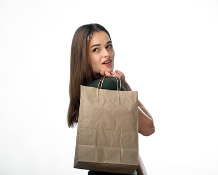 Young female is holding grocery shopping bag. Stands on white background. Woman looking behind back. Young girl is holding light brown shopping bag behind back. Isolated background. Closeup. photo