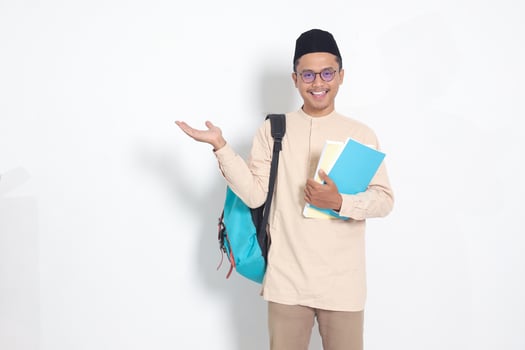 Portrait of excited student Asian muslim man in koko shirt with skullcap carrying backpack, holding school books, pointing to the side. Islamic education concept. Isolated image on white background photo