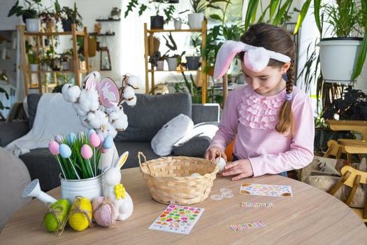 A cute girl with pink bunny ears makes an Easter craft - decorates an egg in the form of a unicorn with rhinestones, horn, flowers in the interior of a house with plants. photo