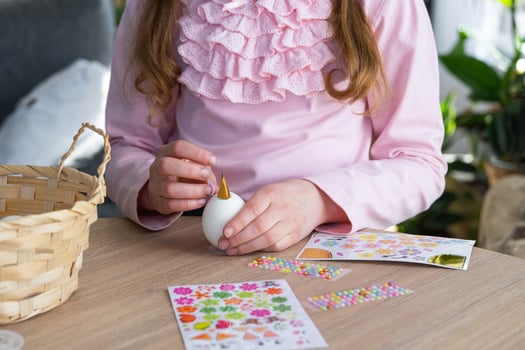 A cute girl with pink bunny ears makes an Easter craft - decorates an egg in the form of a unicorn with rhinestones, horn, flowers in the interior of a house with plants. photo