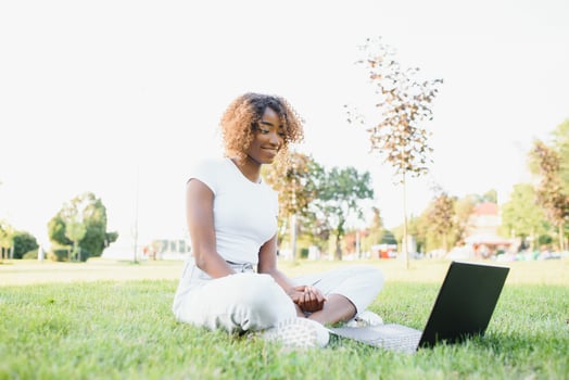 Young attractive dark-skinned college student wearing tank top and jeans sitting on the lawn at campus on sunny day, working on her thesis using laptop computer, looking busy and concentrated photo