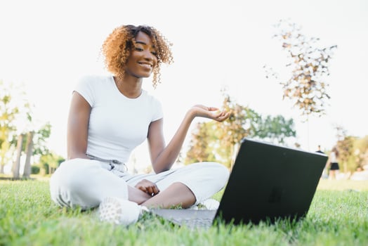 Young smiling black woman sitting outdoors on grass with laptop, typing, surfing internet. Technology, communication, education and remote working concept, copy space photo