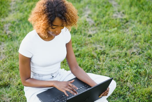 Human and technology concept. Charming African woman with short pixie hairstyle enjoying sunny weather, sitting on the lawn in front of laptop computer in the public garden, waiting for her friend photo