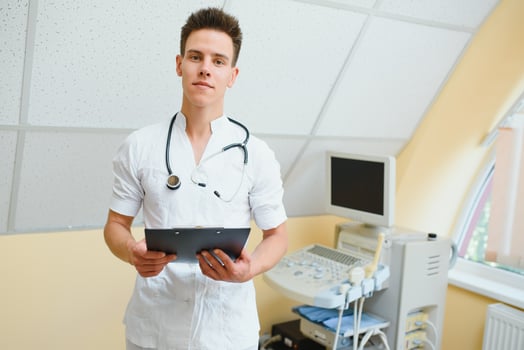 Charming young male doctor , sitting at his office near ultrasound scanning machine. Handsome friendly doctor enjoying working at the hospital, copy space. Ultrasonic concept photo