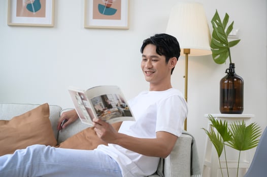 Happy asian man sitting on couch  reading a book in living room at home, relax time and lifestyle concept photo