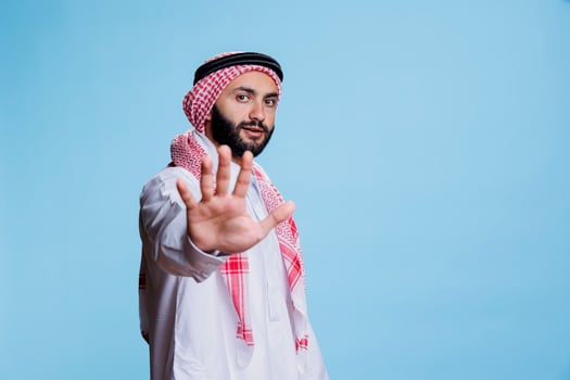 Muslim man wearing traditional clothes showing stop gesture with hand and looking at camera with serious expression. Confident arab posing with prohibition sign studio portrait photo
