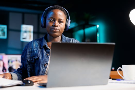 Focused black woman wearing wireless headphones using laptop and writing notes. African american female student with headset watching online webinar, listening to audio course on personal computer. photo