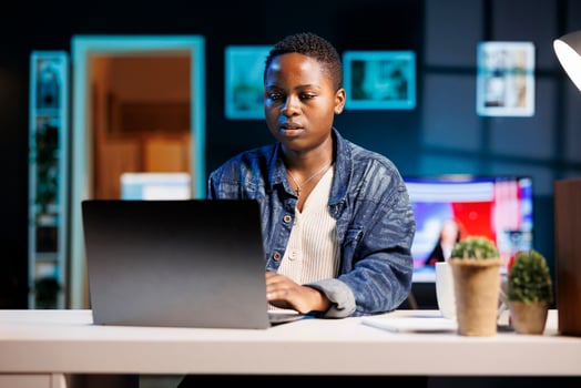 Busy black female freelancer sitting in her modern living room office, working on personal computer. African american college student typing and reviewing information on digital laptop. photo