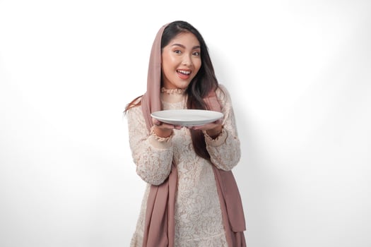 Excited Asian Muslim woman holding plate and cutlery to eat after fasting while smiling cheerfully on isolated white background. Ramadan concept photo