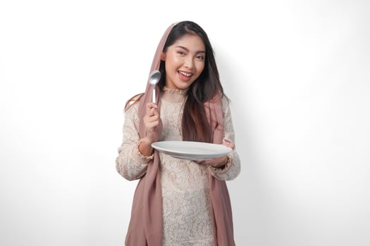 Excited Asian Muslim woman holding plate and cutlery to eat after fasting while smiling cheerfully on isolated white background. Ramadan concept photo