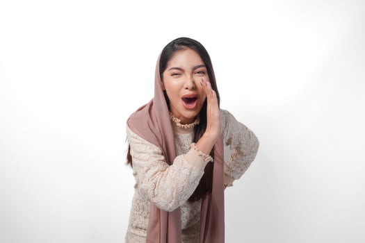 Young Asian Woman wearing headscarf veil hijab is raising hands and shouting with mouth wide open, isolated white background photo