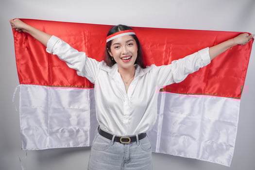 Smiling Asian woman wearing flag headband and casual outfit while holding Indonesia flag at the back. Independence day advertisement concept photo