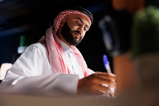 Detailed image showcasing focused Arab entrepreneur at the table writing on a notebook. Close-up shot of a Muslim man using a pen for taking research notes in his home office. photo