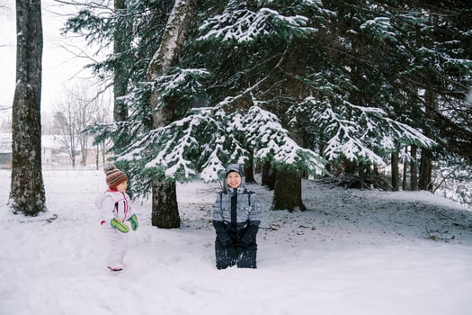 Little girl looks at her smiling mother sitting under a snow-covered spruce tree in a coniferous forest photo