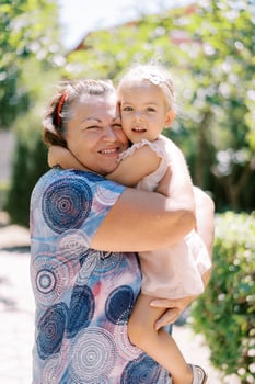 Little smiling girl sits in the arms of her grandmother hugging her in the garden photo