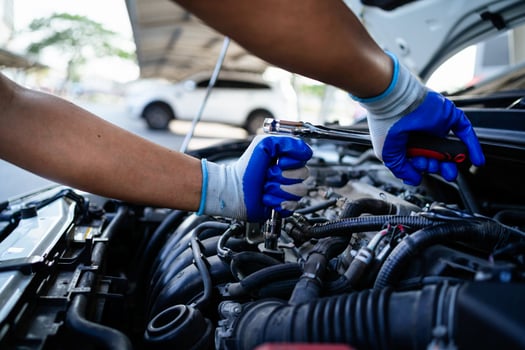 A mechanic is working on a car engine, wearing blue gloves. The car is parked next to a white car photo