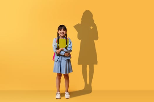 A cheerful young girl clasps a book and smiles, wearing a blue dress and backpack photo