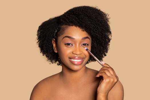 Radiant black woman applying concealer, makeup brush in hand photo