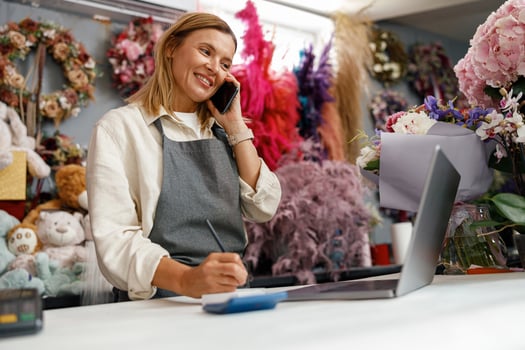 Smiling flower shop owner using her telephone and laptop to take delivery service for her store photo