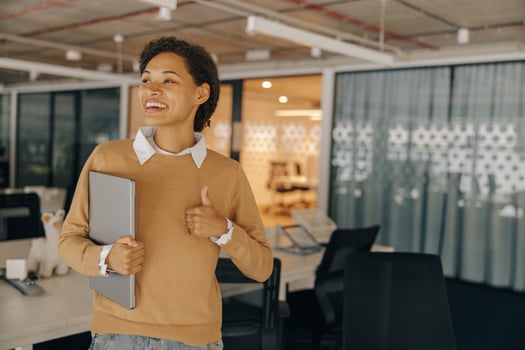 Female manager with laptop standing on background of modern office and showing thumb up photo