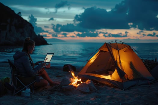 Evening tent camping in seashore. Female traveler uses laptop computer sitting by campfire. Woman on remote work photo