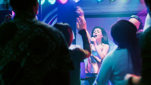 Confident woman doing karaoke on stage, singing songs with male DJ in front of people at nightclub. Happy girl having fun with live perfromance next to audio mixing station in club. photo