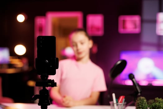 Focus on smartphone on tripod used by little girl shooting product unboxing for streaming channel on online platforms. Child in blurry background warmly welcoming fans, filming reviews photo