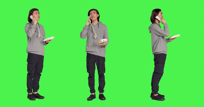 Asian guy talking on landline phone, answering call on telephone with cord while he stands against full body greenscreen. Young person using retro office phone for remote communication. photo
