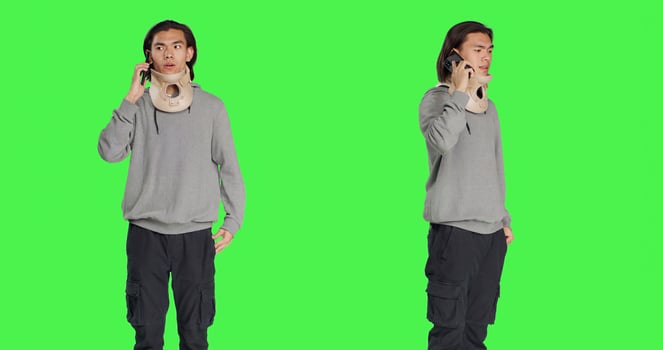 Young adult with neck collar answers call on smartphone, talking to people about his bone fracture accident. Hurt person wearing cervical foam brace discussing on telephone line, greenscreen. photo