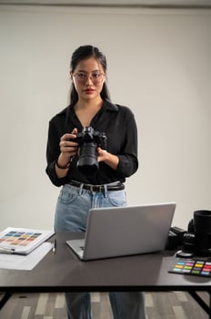 An elegant Asian female photographer in a black shirt with her DSLR camera is standing in her studio photo