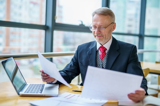 Mature businessman looking at laptop reading and printing improvements sitting near the window in his office. Panoramic city view background. Business photo. photo