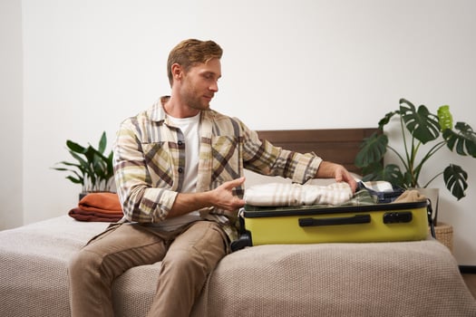 Portrait of young man packing his luggage for a trip, going on holiday, sitting on bed and putting clothes in suitcase, preparing for his flight photo