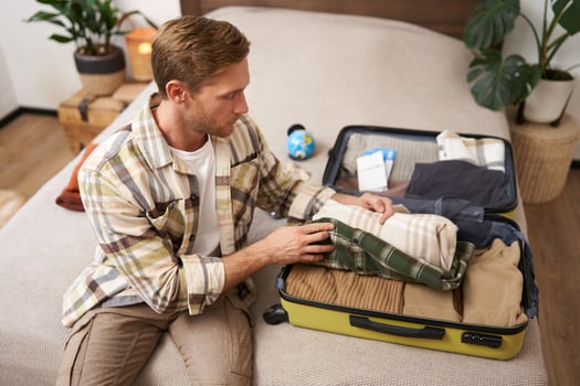 Portrait of young man sits on bed with suitcase, goes on holiday, packs clothes for vacation or business trip, checks his luggage photo